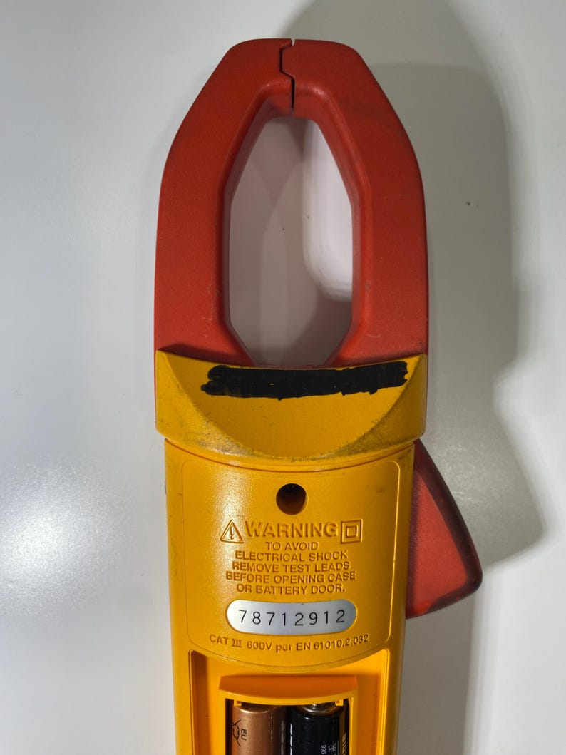 Pu&ograve; includere: Una pinza amperometrica gialla e rossa con un'etichetta di avvertenza che recita: WARNING TO AVOID ELECTRICAL SHOCK REMOVE TEST LEADS BEFORE OPENING CASE OR BATTERY DOOR. Il dispositivo include il numero di serie 78712912 e CAT III 600V per EN 61010.2.032.