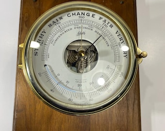 May include: A wall-mounted barometer featuring a circular brass frame and a white dial with black text reading "STORMY," "RAIN," "CHANGE," "FAIR," and "VERY DRY." The device includes a thermometer with Celsius and Fahrenheit scales, all mounted on a rectangular wooden board.