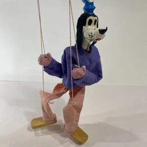 May include: A Goofy marionette puppet featuring a blue hat, a purple long-sleeved shirt, and light pink pants. The character has black ears, a white muzzle, and is suspended by thin strings attached to wooden feet and hands for movement.