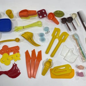 May include: A selection of kitchen utensils and tools in various colours. Includes measuring cups, spoons, spatulas, knives, combs, and other items. The items are made of plastic and are arranged on a white surface.
