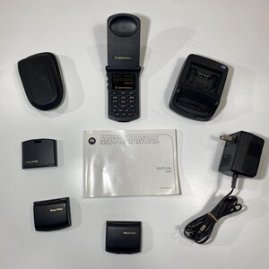 Puede incluir: Un tel&eacute;fono plegable Motorola Razr V3c negro, una base de carga, un adaptador de corriente con cable, tres bater&iacute;as negras, un estuche de transporte negro y un folleto Motomanual blanco. El tel&eacute;fono est&aacute; abierto, mostrando un teclado y pantalla.