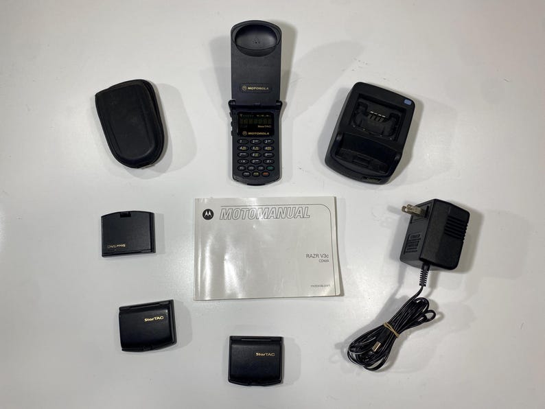 Puede incluir: Un tel&eacute;fono plegable Motorola Razr V3c negro, una base de carga, un adaptador de corriente con cable, tres bater&iacute;as negras, un estuche de transporte negro y un folleto Motomanual blanco. El tel&eacute;fono est&aacute; abierto, mostrando un teclado y pantalla.