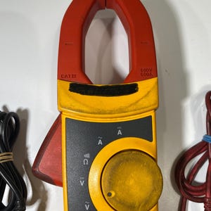 Pu&ograve; includere: Una pinza amperometrica digitale gialla e rossa dotata di un grande quadrante circolare, un grilletto rosso laterale e una ganascia superiore. Il dispositivo presenta marcature tra cui CAT III, 600V, 600A e vari simboli elettrici di misurazione.