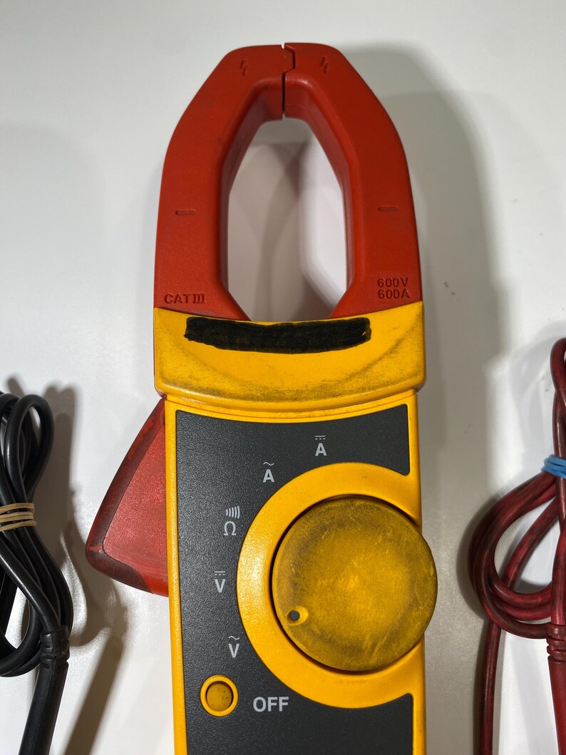 Pu&ograve; includere: Una pinza amperometrica digitale gialla e rossa dotata di un grande quadrante circolare, un grilletto rosso laterale e una ganascia superiore. Il dispositivo presenta marcature tra cui CAT III, 600V, 600A e vari simboli elettrici di misurazione.