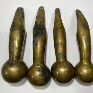 Könnte beinhalten: Vier antike Möbelbeine aus Messing, jedes mit einem abgerundeten Sockel und einer langen, spitz zulaufenden Oberseite. Das Messing hat ein gealtertes, angelaufenes Aussehen mit dunkleren Flecken. Jedes Bein hat oben ein kleines Loch. Die Beine sind in einer Reihe angeordnet.