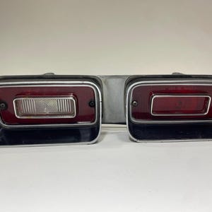 May include: A dual automotive taillight assembly featuring two rectangular housings with black trim and silver borders. The left unit contains a clear centre lens over a red background, while the right unit features a solid red lens.