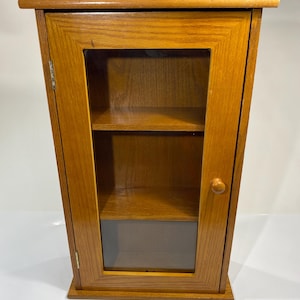 May include: A wall-mounted wooden cabinet with a natural oak finish, featuring a single hinged door with a glass panel. The cabinet includes two interior shelves and a small round wooden knob for opening the door, showcasing a simple rectangular frame.