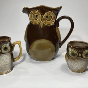 May include: A ceramic owl-themed pitcher and two mugs. The pitcher is dark brown with a large handle and a spout. The mugs and pitcher have owl faces with large eyes and are in shades of brown, tan, and cream.