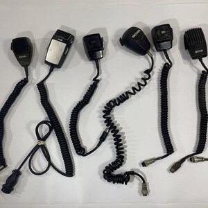 May include: Eight black handheld microphones with coiled cords. The microphones have a variety of shapes and sizes.