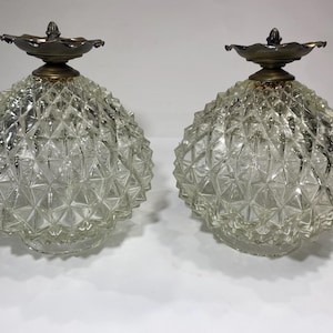 May include: A pair of spherical clear glass light shades featuring a textured diamond-cut pattern. Each shade is topped with a dark metallic brass-toned finial cap with a scalloped edge, providing a decorative finish to the glass globes.