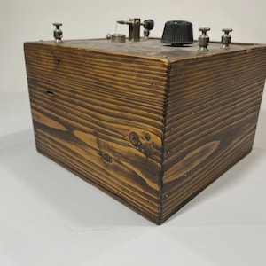 May include: A vintage wooden box with a dark stain, featuring metal components on top, including knobs and posts. The box has a rectangular shape with visible wood grain and is likely a piece of antique equipment.