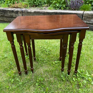 Peut inclure: Un ensemble de deux tables gigognes avec une finition en bois brun foncé. Chaque table présente un plateau rectangulaire avec un bord avant légèrement incurvé et quatre pieds fins et cannelés. Les tables sont conçues pour s'emboîter, la plus petite se glissant sous la plus grande, révélant un grain de bois sombre et uniforme sur toute la structure.