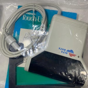 May include: An ALFA DATA touch-up device with a gray body and black base, packaged in plastic. The device is connected to a gray cable. A blue and green booklet with the text "MIGRAPH Touch-Up" is also visible.
