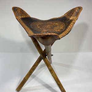 May include: A folding stool with a dark brown leather seat featuring an embossed design. The seat is supported by bamboo legs. The stool is designed for portability and outdoor use.