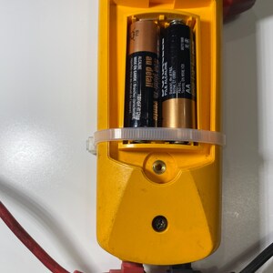 Pu&ograve; includere: Un multimetro elettrico giallo con vano batteria aperto contenente due batterie AA. Il dispositivo presenta il numero di serie 78712912, una classificazione CAT III 600V ed &egrave; collegato a un cavo di test elettrico rosso e uno nero angolati.