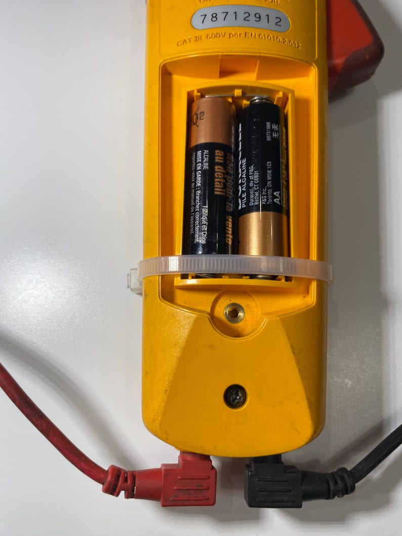 Pu&ograve; includere: Un multimetro elettrico giallo con vano batteria aperto contenente due batterie AA. Il dispositivo presenta il numero di serie 78712912, una classificazione CAT III 600V ed &egrave; collegato a un cavo di test elettrico rosso e uno nero angolati.
