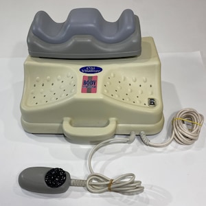 Könnte beinhalten: Ein beigefarbenes Chi Vitalizer Body Stimulator Gerät mit einer grauen, konturierten Fuß- oder Knöchelauflage auf der Oberseite. Das Gerät verfügt über einen zentralen Griff, einen Netzschalter und eine kabelgebundene Fernbedienung mit schwarzem Drehregler.