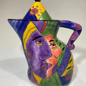 May include: A ceramic pitcher with a lid featuring a vibrant, multi-colored abstract face design. The surface displays bold geometric shapes in purple, orange, yellow, green, and blue, with stylized facial features including eyes and lips.