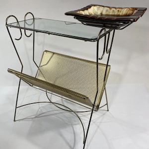 May include: A metal magazine rack with a gold-tone finish, featuring a glass top shelf and a perforated metal basket. A square, brown and cream ceramic dish with a glossy glaze is positioned on the upper right side of the metal frame structure.