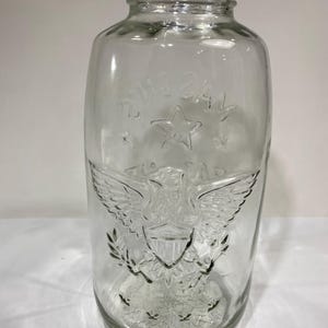 May include: A clear glass jar with a threaded neck featuring an embossed design of an eagle with spread wings, a shield on its chest, and a star above it. The glass surface displays moulded details including laurel branches and a textured base.