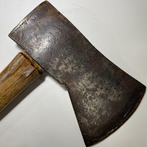 May include: A metal axe head with a weathered, dark gray finish and visible rust spots, featuring the stamped text 3 1/2 on the surface. The axe head is attached to a light brown wooden handle with a visible grain pattern and a worn, textured finish.