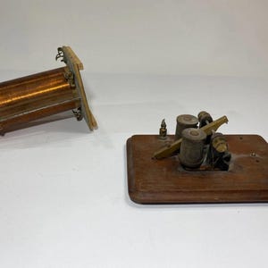 May include: A vintage electrical component featuring a cylindrical copper wire coil mounted between two wooden end plates, positioned next to a telegraph key mechanism with two metal spools mounted on a rectangular wooden base plate.