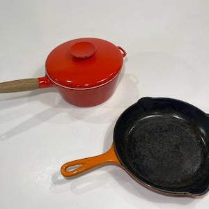May include: A bright red enameled saucepan with a wooden handle and a matching lid, positioned next to a black cast iron skillet with an orange exterior handle. Both pieces of cookware feature long handles and are designed for stovetop use.