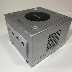 May include: Silver GameCube console with a black disc tray cover. The console has multiple controller ports and a ventilation grid on the side. The GameCube logo is visible on the top.
