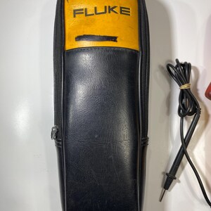 Pu&ograve; includere: Una custodia con cerniera nera e gialla con la scritta FLUKE stampata in nero sulla sezione gialla. Accanto alla custodia si trova un puntale elettrico nero con punta metallica e filo avvolto fissato con un elastico.
