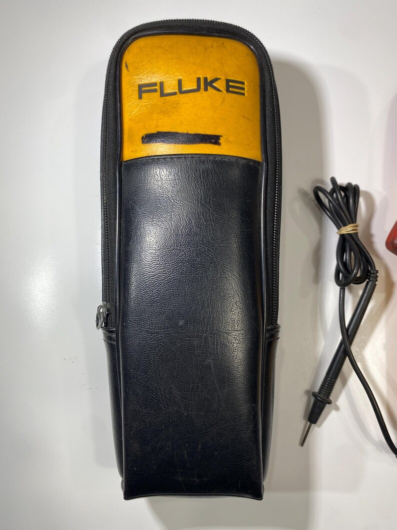Pu&ograve; includere: Una custodia con cerniera nera e gialla con la scritta FLUKE stampata in nero sulla sezione gialla. Accanto alla custodia si trova un puntale elettrico nero con punta metallica e filo avvolto fissato con un elastico.