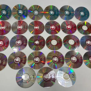 May include: A collection of twenty-five circular compact discs arranged in five rows. Each disc features a reflective, iridescent surface with varying colour patterns and printed text labels, including titles like Management and various numbered indicators.