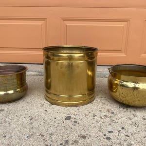 May include: Three polished brass containers of varying shapes and sizes. The left container is a short, wide bowl with horizontal ridges. The centre container is a tall, smooth cylinder. The right container is a rounded pot with a handle attachment.