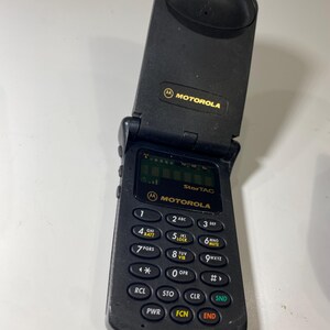 Puede incluir: Un tel&eacute;fono m&oacute;vil plegable Motorola StarTAC de color negro con dise&ntilde;o vertical. El dispositivo cuenta con una peque&ntilde;a pantalla digital, un teclado num&eacute;rico con botones circulares y el logotipo de Motorola impreso en dorado en la parte superior e inferior.