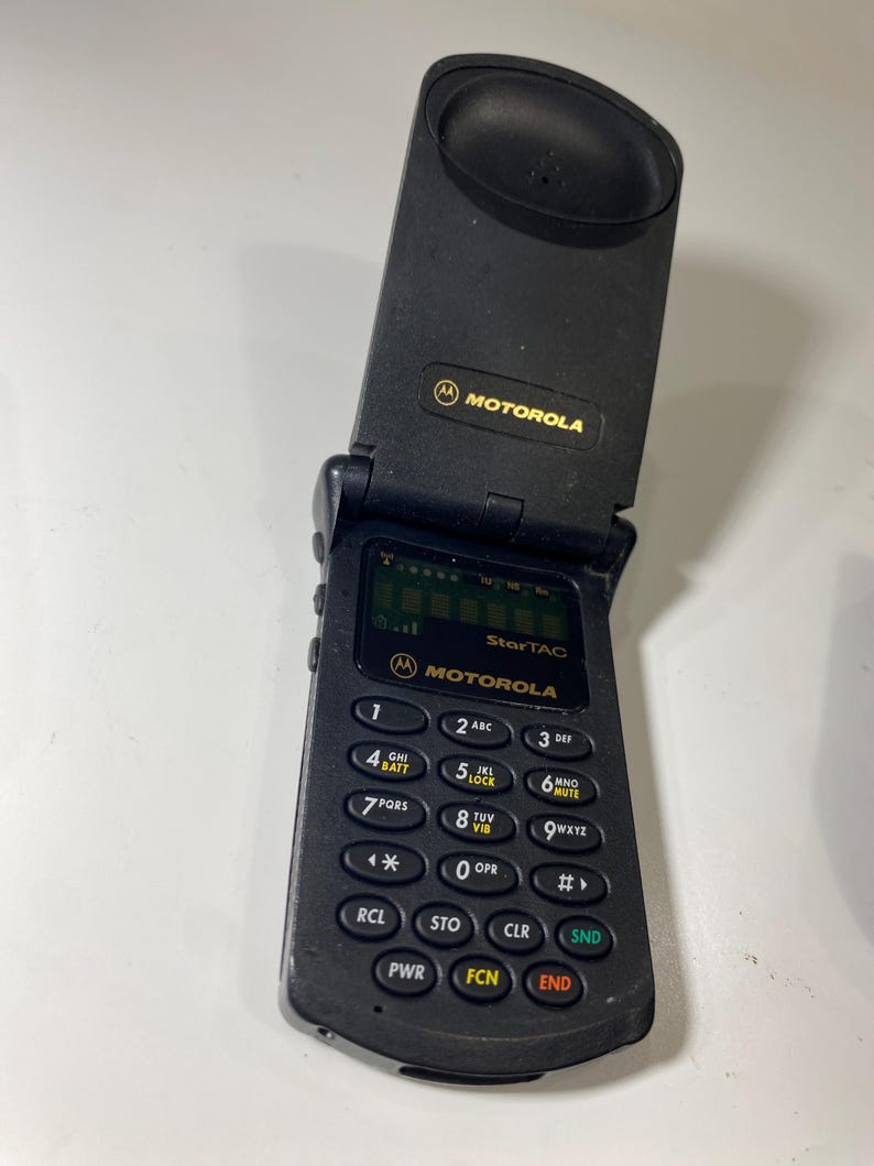 Puede incluir: Un tel&eacute;fono m&oacute;vil plegable Motorola StarTAC de color negro con dise&ntilde;o vertical. El dispositivo cuenta con una peque&ntilde;a pantalla digital, un teclado num&eacute;rico con botones circulares y el logotipo de Motorola impreso en dorado en la parte superior e inferior.