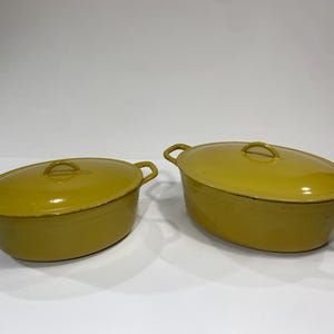 May include: Two oval-shaped, mustard yellow enamelled cast iron Dutch ovens with matching lids. Each pot features two side handles and a central lid handle. The exterior finish is smooth and glossy, showing a consistent mustard yellow colour.