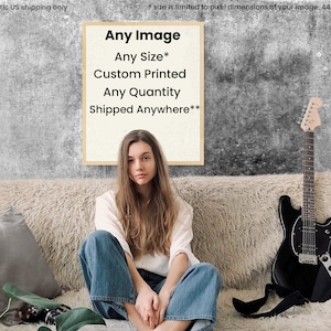 May include: A framed poster with the text "Any Image", "Any Size*", "Custom Printed", "Any Quantity", and "Shipped Anywhere**" on a white background. The poster is hanging on a gray wall behind a couch with a woman sitting on it. A black electric guitar is leaning against the couch.