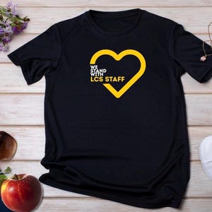 Puede incluir: Camiseta negra con un corazón amarillo y el texto "WE STAND WITH LCS STAFF". La camiseta está sobre una superficie de madera con una biblia, gafas de sol y una manzana. Otros artículos están presentes.