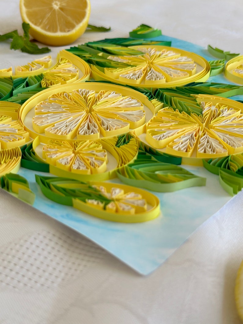 Puede incluir: Una obra de arte de quilling de papel con rodajas de lim&oacute;n y hojas. Los limones son amarillos y blancos, con hojas verdes. Hay medio lim&oacute;n en el fondo. La obra de arte est&aacute; sobre un fondo azul claro.