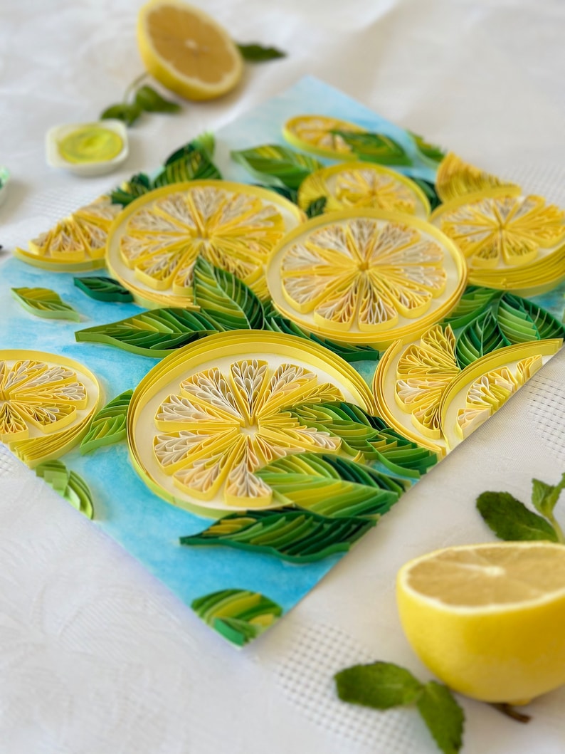 Puede incluir: Una obra de arte de quilling de papel hecha a mano con rodajas de lim&oacute;n y hojas verdes sobre un fondo azul claro. Los limones son amarillos con centros blancos, y las hojas son de varios tonos de verde. Tambi&eacute;n hay medio lim&oacute;n y hojas de menta.
