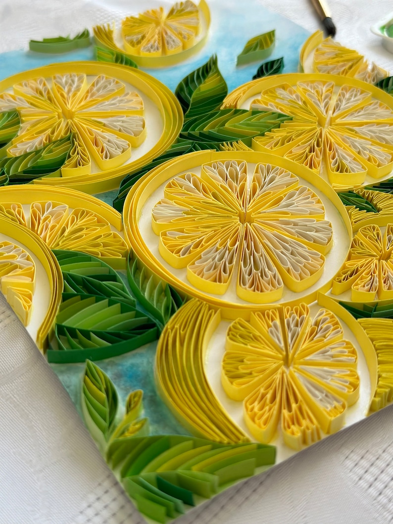 Puede incluir: Una obra de arte de quilling de papel hecha a mano que presenta rodajas de lim&oacute;n y hojas verdes sobre un fondo azul claro. Los limones son amarillos y blancos, con dise&ntilde;os detallados de espirales de papel. Vista de cerca.