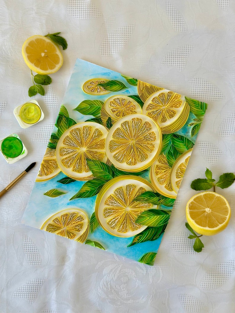 Puede incluir: Una pintura a la acuarela con rodajas de lim&oacute;n y hojas verdes sobre un fondo azul claro. Medias rodajas de lim&oacute;n fresco y ramitas de menta est&aacute;n dispuestas alrededor de la pintura. Tambi&eacute;n se ven pinturas de acuarela y un pincel.