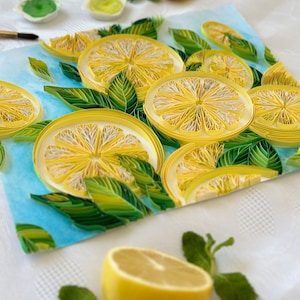 Puede incluir: Una vibrante obra de arte de quilling de papel que presenta rodajas de lim&oacute;n y hojas verdes sobre un fondo azul claro. La obra de arte incluye medio lim&oacute;n y ramitas de menta. Los limones son amarillos con centros blancos, y las hojas son verdes.