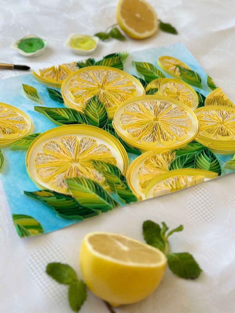 Puede incluir: Una vibrante obra de arte de quilling de papel que presenta rodajas de lim&oacute;n y hojas verdes sobre un fondo azul claro. La obra de arte incluye medio lim&oacute;n y ramitas de menta. Los limones son amarillos con centros blancos, y las hojas son verdes.
