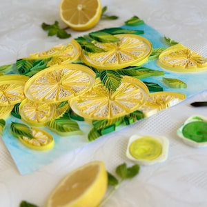 Puede incluir: Naturaleza muerta con una obra de arte de quilling de rodajas de lim&oacute;n y hojas, con medio lim&oacute;n y ramitas de menta. La obra utiliza papel amarillo, blanco y verde sobre un fondo azul claro. La escena est&aacute; sobre una superficie blanca texturizada.