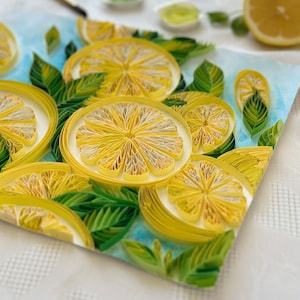 Puede incluir: Primer plano de una obra de arte de quilling de papel con rodajas de lim&oacute;n amarillas y hojas verdes sobre un fondo azul. Los limones est&aacute;n hechos de tiras de papel enrolladas, con medio lim&oacute;n y materiales de pintura en el fondo. La obra est&aacute; sobre una superficie blanca.