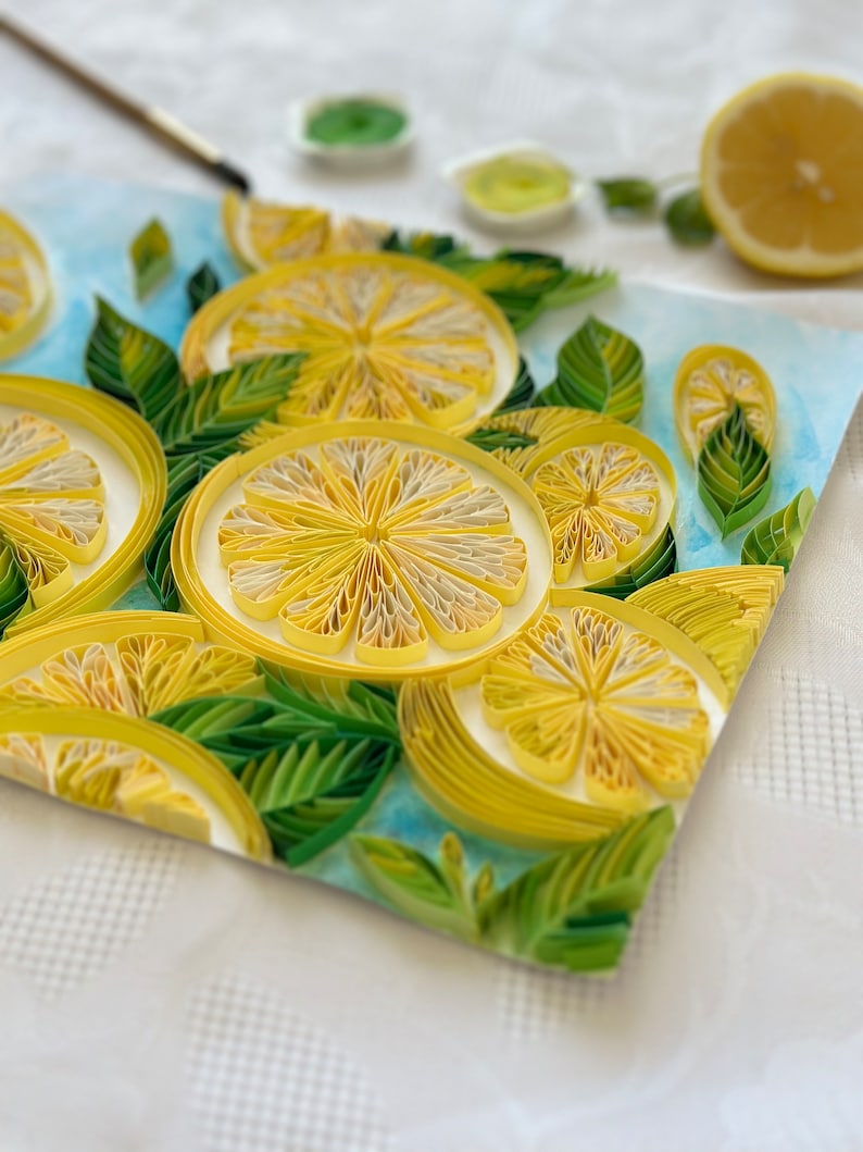 Puede incluir: Primer plano de una obra de arte de quilling de papel con rodajas de lim&oacute;n amarillas y hojas verdes sobre un fondo azul. Los limones est&aacute;n hechos de tiras de papel enrolladas, con medio lim&oacute;n y materiales de pintura en el fondo. La obra est&aacute; sobre una superficie blanca.