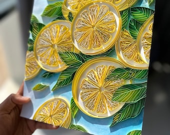 May include: A paper quilling art piece featuring sliced lemons and green leaves against a light blue background. The lemons are yellow with white details, and the leaves are various shades of green. The artwork is held by a person.