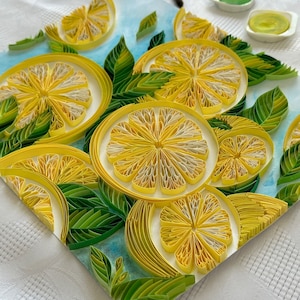 Puede incluir: Una detallada obra de arte de quilling de papel con rodajas de lim&oacute;n y hojas verdes. Los limones son amarillos con centros blancos, y las hojas son de varios tonos de verde. Un pincel y pintura son visibles en el fondo.