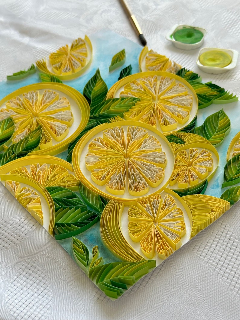 Puede incluir: Una detallada obra de arte de quilling de papel con rodajas de lim&oacute;n y hojas verdes. Los limones son amarillos con centros blancos, y las hojas son de varios tonos de verde. Un pincel y pintura son visibles en el fondo.
