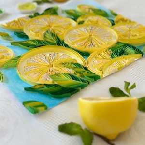 Puede incluir: Primer plano de una obra de arte de quilling de papel con rodajas de lim&oacute;n amarillas con centros blancos y hojas verdes sobre un fondo azul claro. Un lim&oacute;n a la mitad y hojas de menta en primer plano.